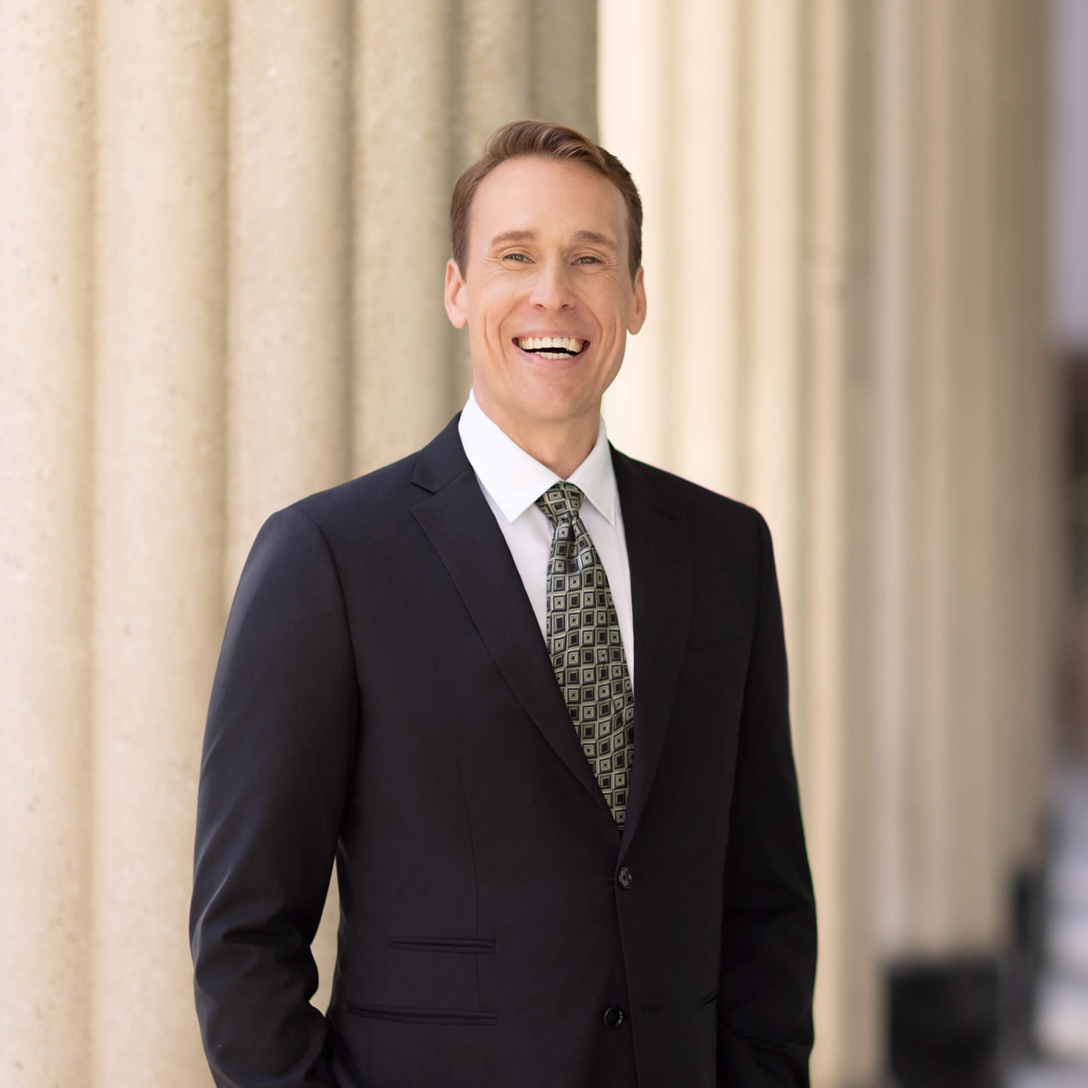Smiling man wearing a black suit and patterned tie standing against beige columns.