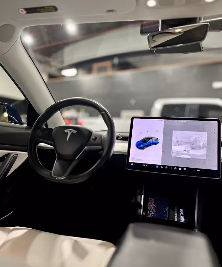 Tesla car interior showing black steering wheel and large touchscreen displaying vehicle images and controls.