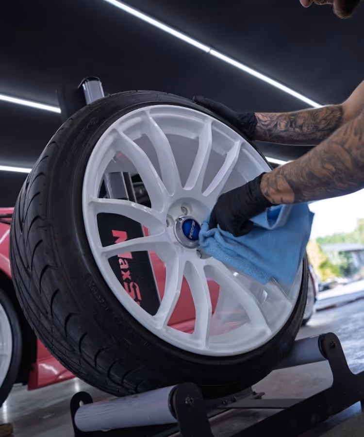 Person with tattooed arms wearing gloves cleans white car wheel rim with blue cloth indoors.