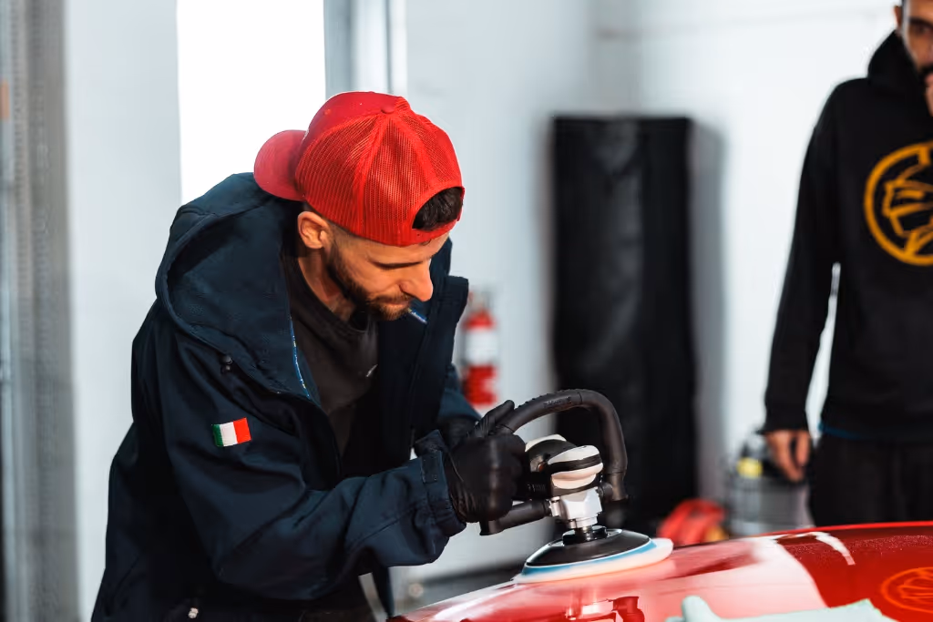 Man in red cap polishing a red car with a power buffer inside a garage.