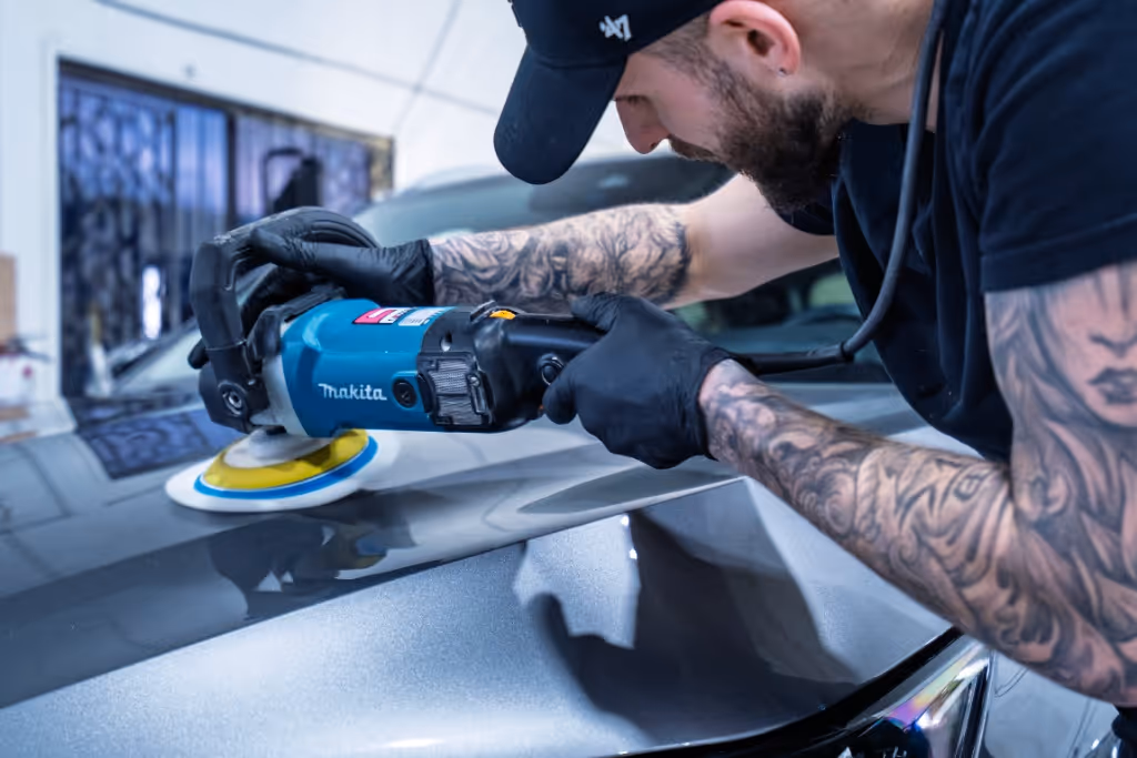 Man with tattooed arms polishing a gray car hood using a blue Makita power buffer.