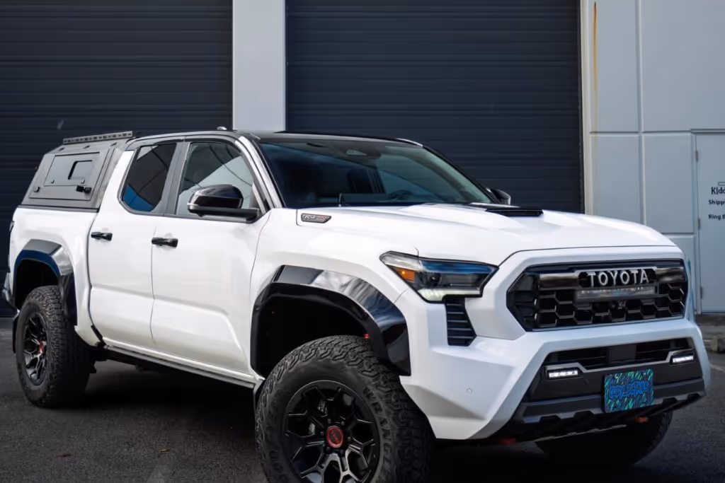 White Toyota pickup truck with large off-road tires and a canopy mounted on the bed parked on pavement.