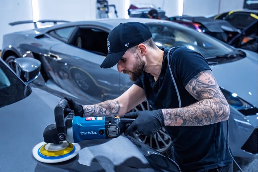 Tattooed man in black using a Makita buffer to polish a grey car in a garage.
