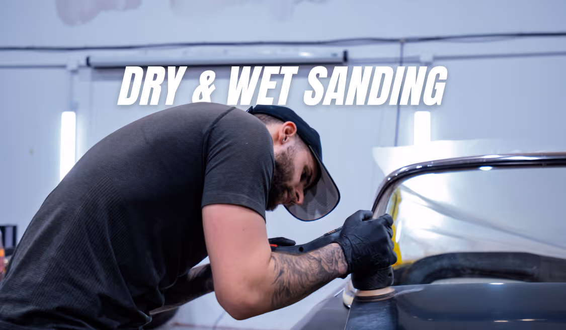 Man wearing black gloves sanding car surface with a power tool in a workshop.
