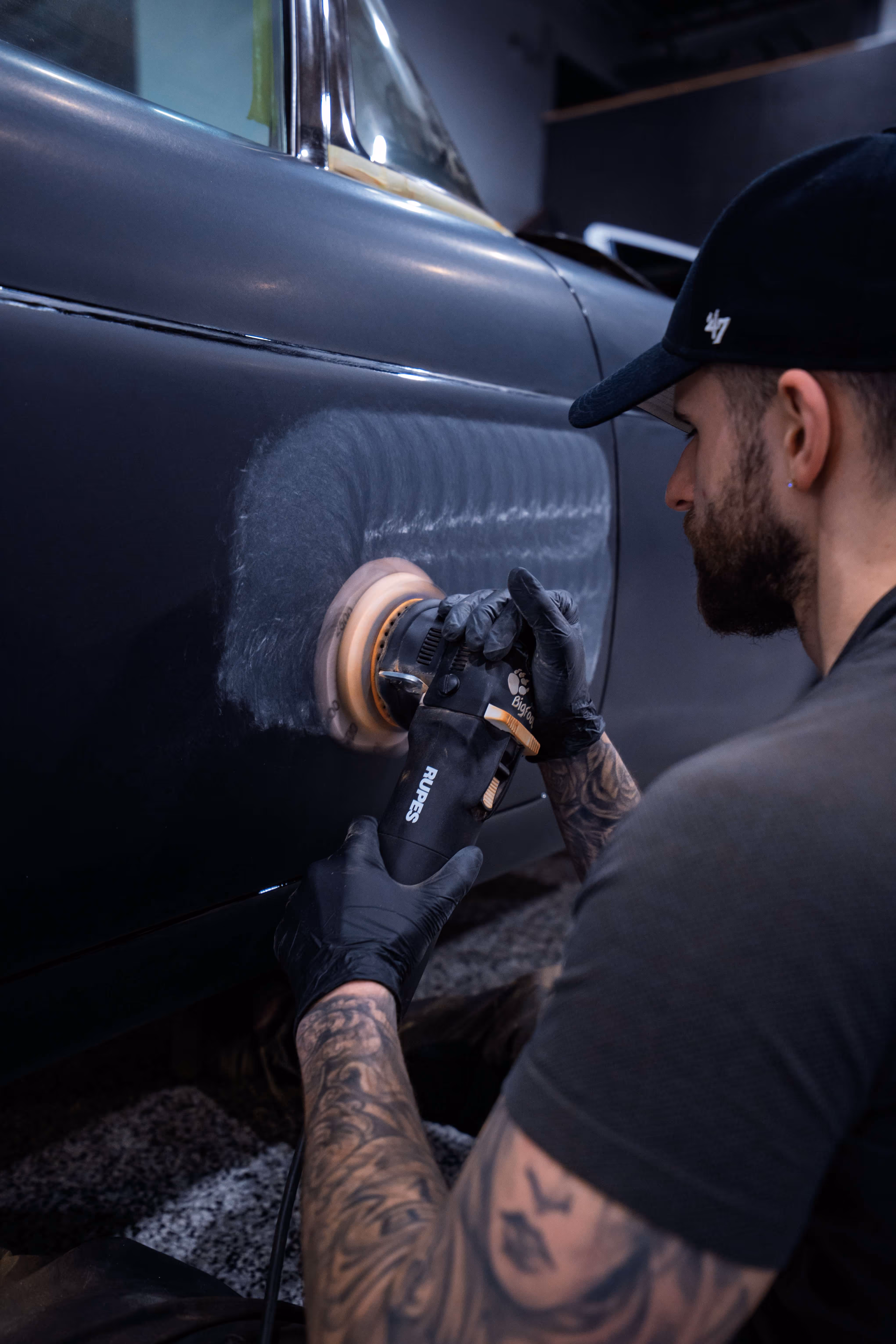 Man with tattoos and gloves polishing a black car door with a Rupes buffer machine.