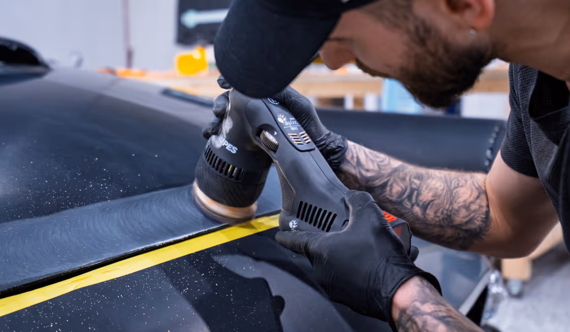 Man with tattoos uses a rotary tool to sand a black car's surface near taped edge.