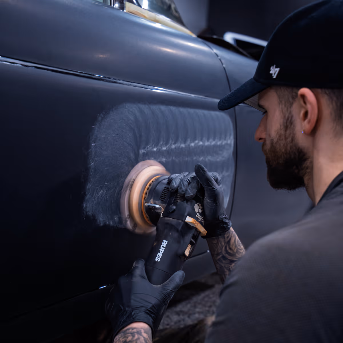 Man in black gloves polishing a black car door using an electric buffer polisher.