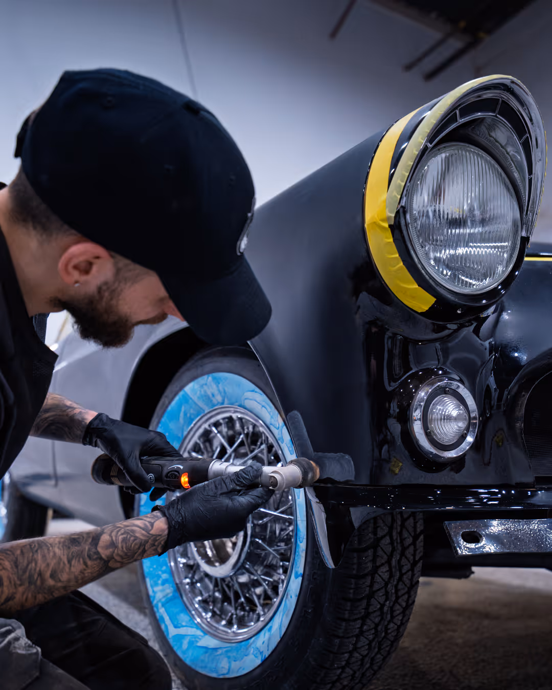 Man wearing black cap and gloves polishes fender of black classic car with power tool.