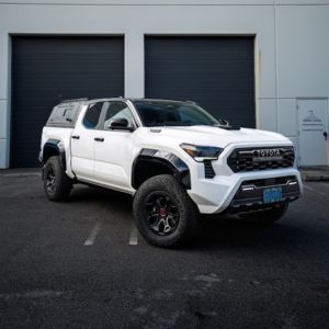 White Toyota pickup truck with off-road tires parked outside a building with black garage doors.