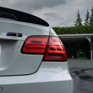 Rear side of white BMW M3 showing taillight and badge with a cloudy sky and greenery background.