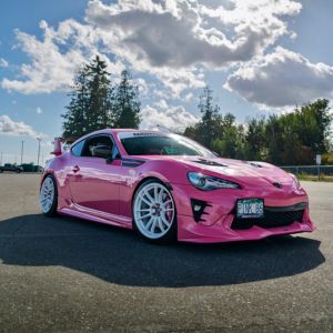 Pink sports car with white wheels parked on asphalt under blue sky with clouds and trees.
