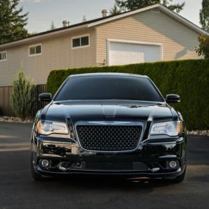 Front view of a shiny black luxury sedan parked on driveway in front of a beige house with hedges.
