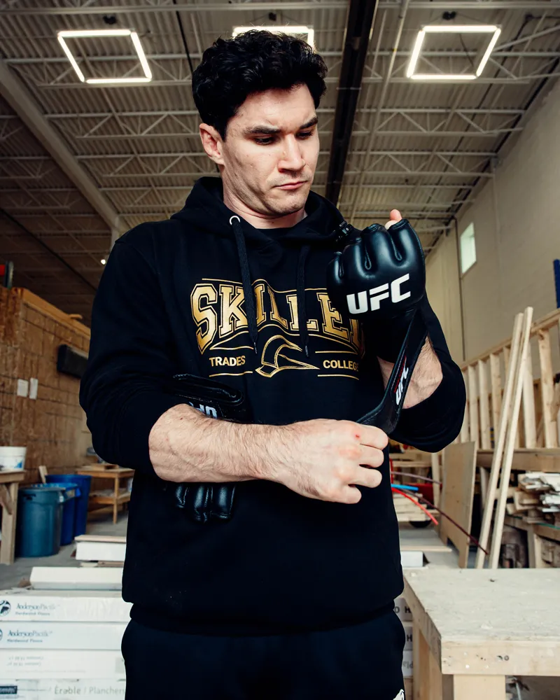 Man wearing a black Skill Trades College hoodie adjusts UFC gloves in a workshop with wood and tools.