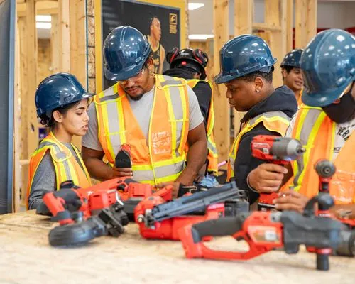 Group of construction workers wearing hard hats and safety vests gathered around a table with various power tools in a workshop setting.