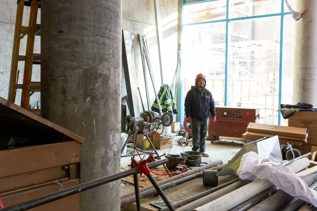 Construction worker wearing a hard hat and jacket standing in a cluttered indoor construction site with pipes, equipment, and large windows in the background.