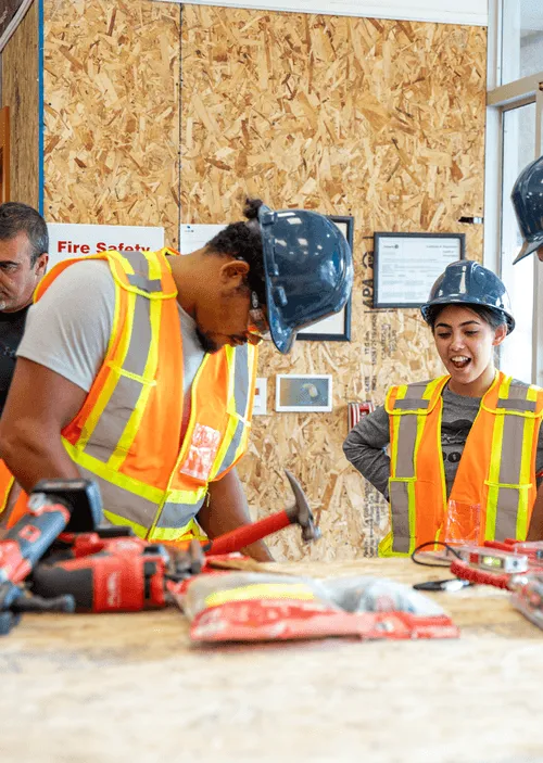 Construction workers wearing safety vests and helmets working with tools on a wooden surface indoors.