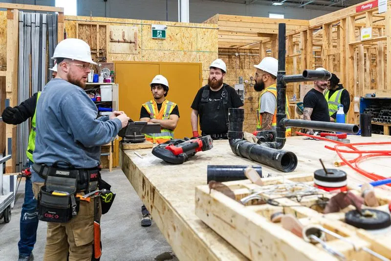 Group of construction workers wearing hard hats and safety vests gathered around a workbench with tools and pipes in an indoor workshop.