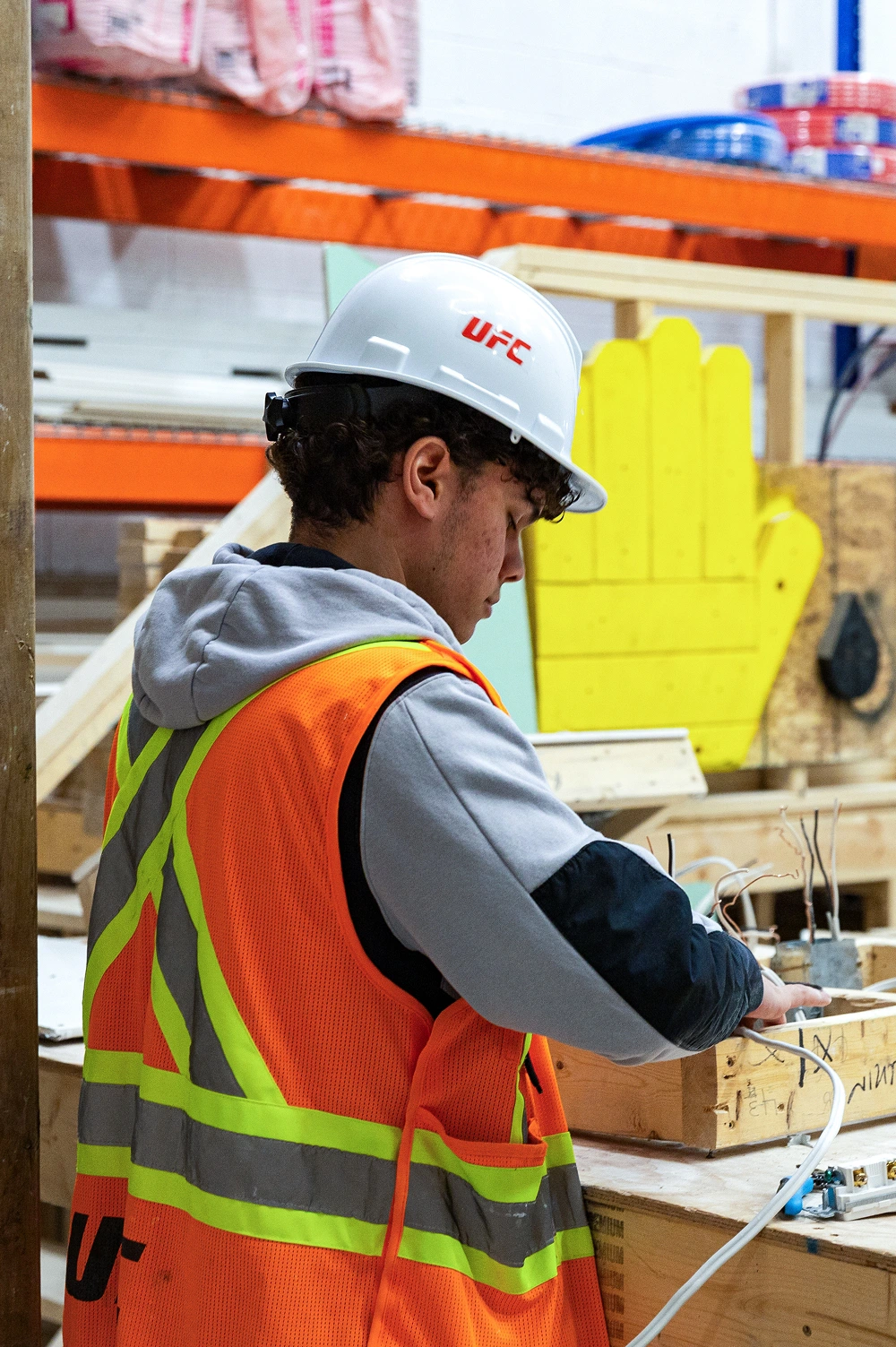 Young man wearing a UFC hard hat and orange safety vest working with electrical wires in a workshop.