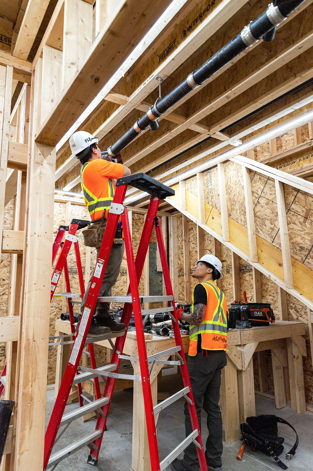 Two construction workers wearing orange safety vests and white helmets installing black pipes in a wooden framed building interior.