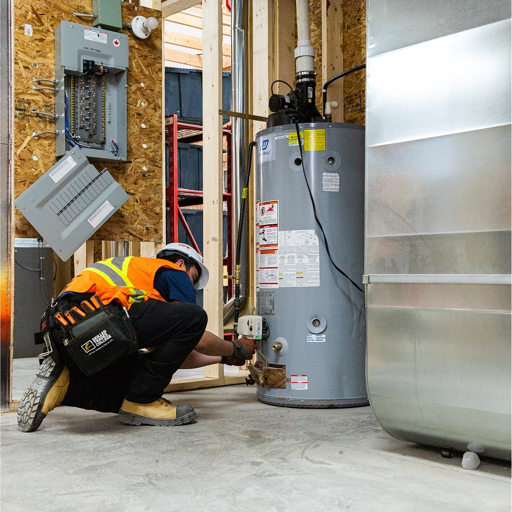 HVAC technician in safety gear crouching to inspect a large gray water heater in a building under construction.