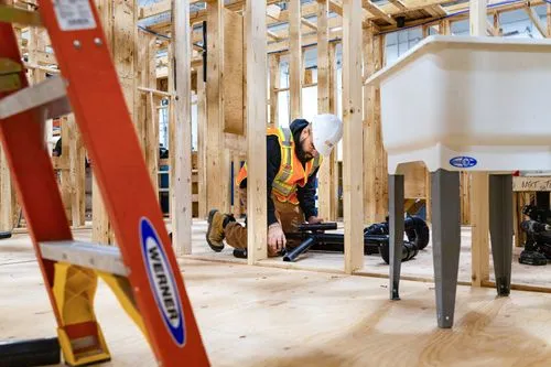 Construction worker wearing a hard hat and safety vest kneeling inside a wooden frame structure, handling black pipes.