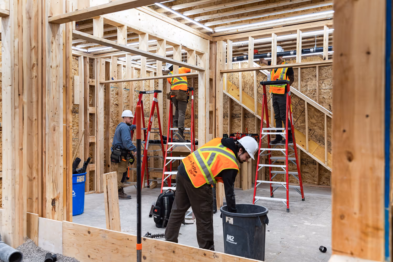 Four construction workers wearing helmets and safety vests working inside a wooden frame building under construction.