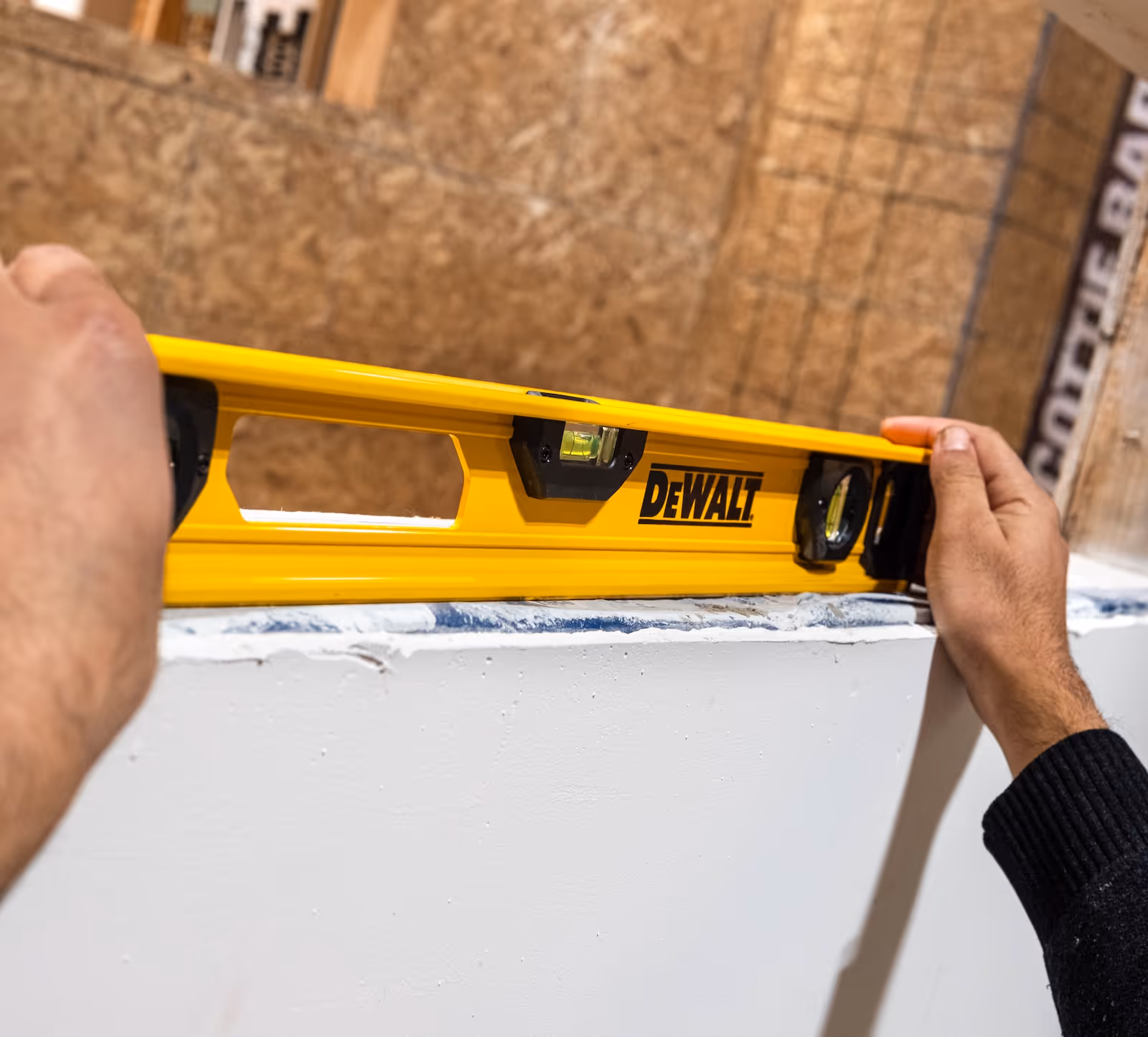 Hands holding a yellow DeWalt spirit level horizontally against a white surface to check its flatness.