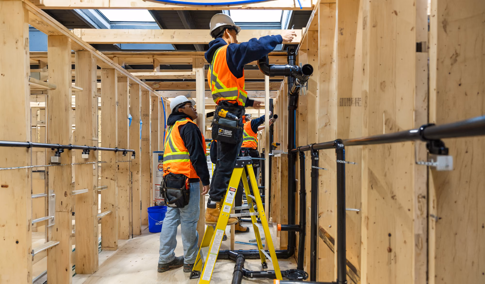 Two construction workers in safety vests and helmets installing black pipes in a wooden framed room.