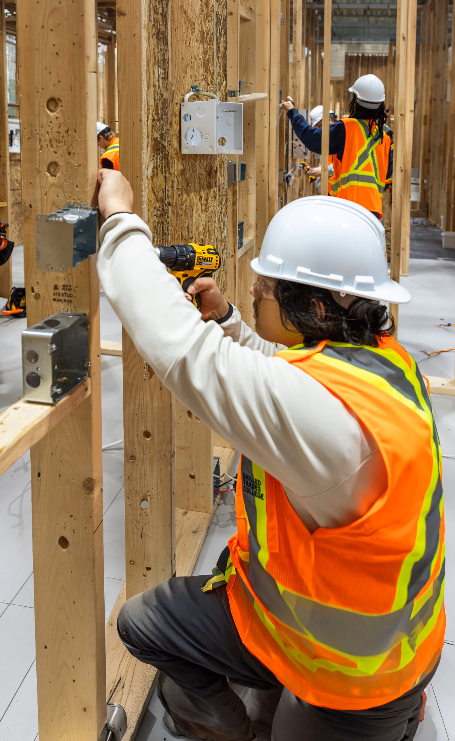 Construction worker in a white helmet and orange safety vest using a drill on wooden framing indoors.