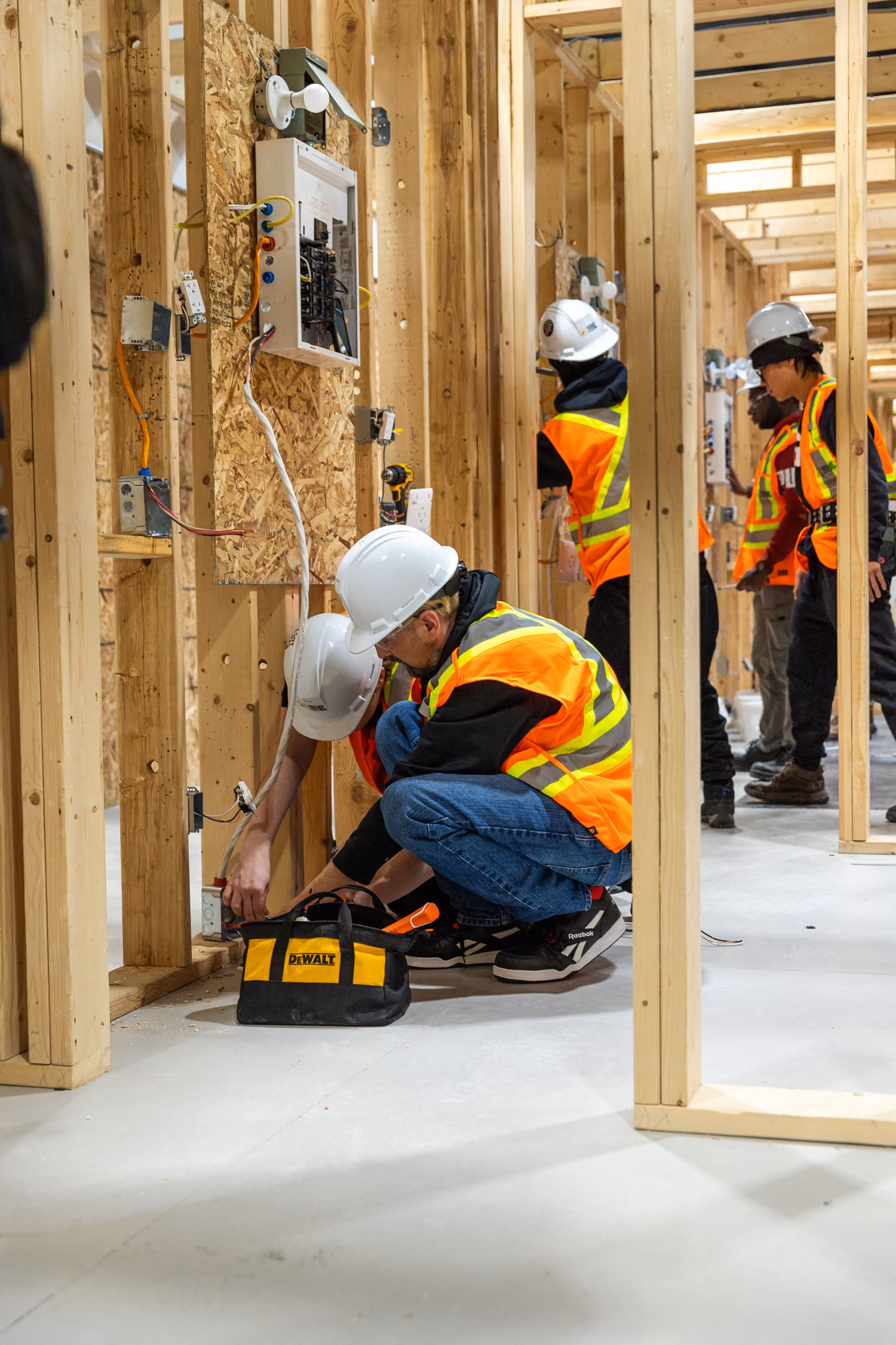 Workers in hard hats and orange vests installing electrical wiring in wooden framed walls indoors.