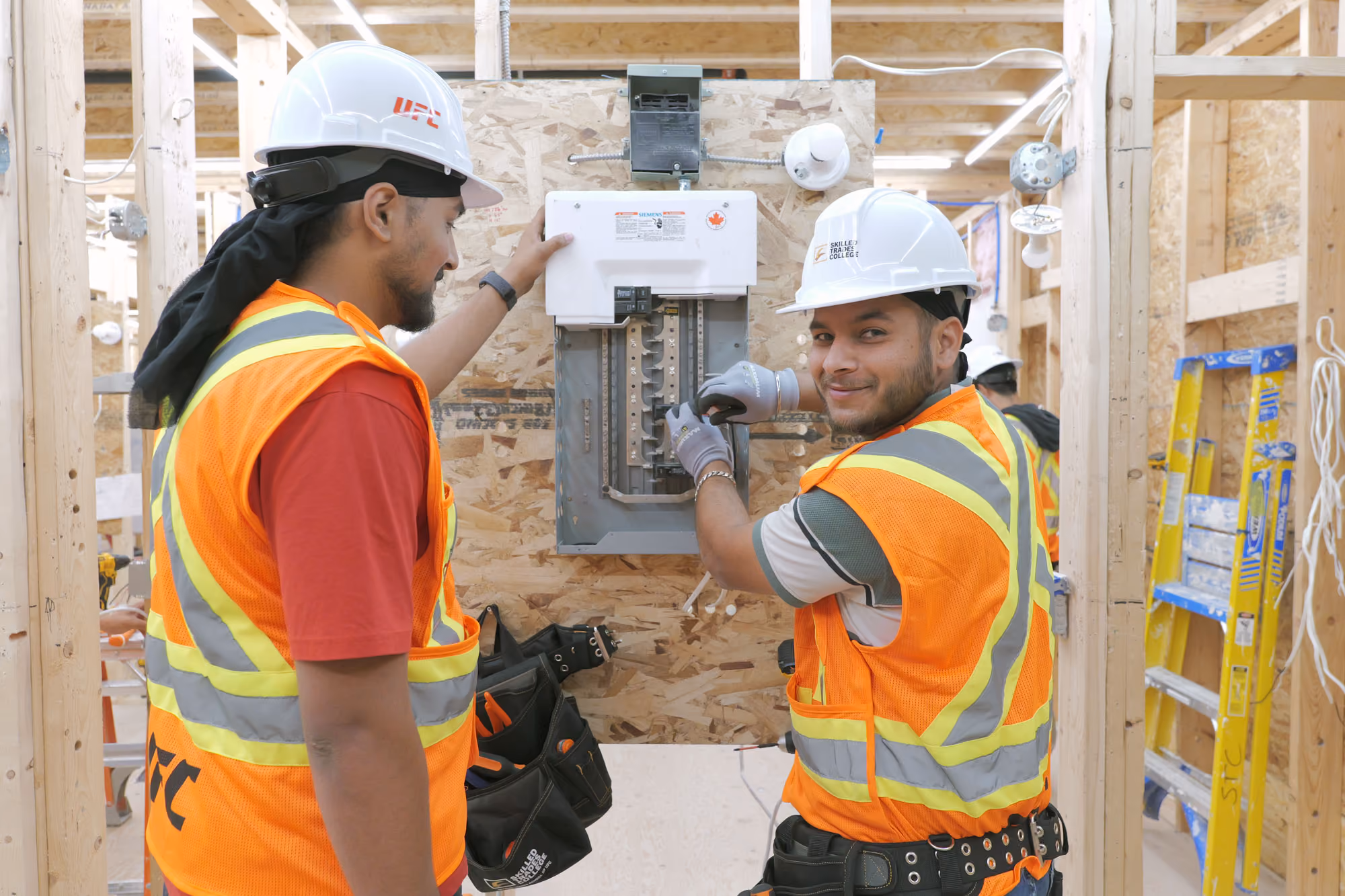 Two construction workers in hard hats and orange safety vests working on an electrical panel mounted on a wooden frame inside a building under construction, with one worker adjusting the panel and the other observing.