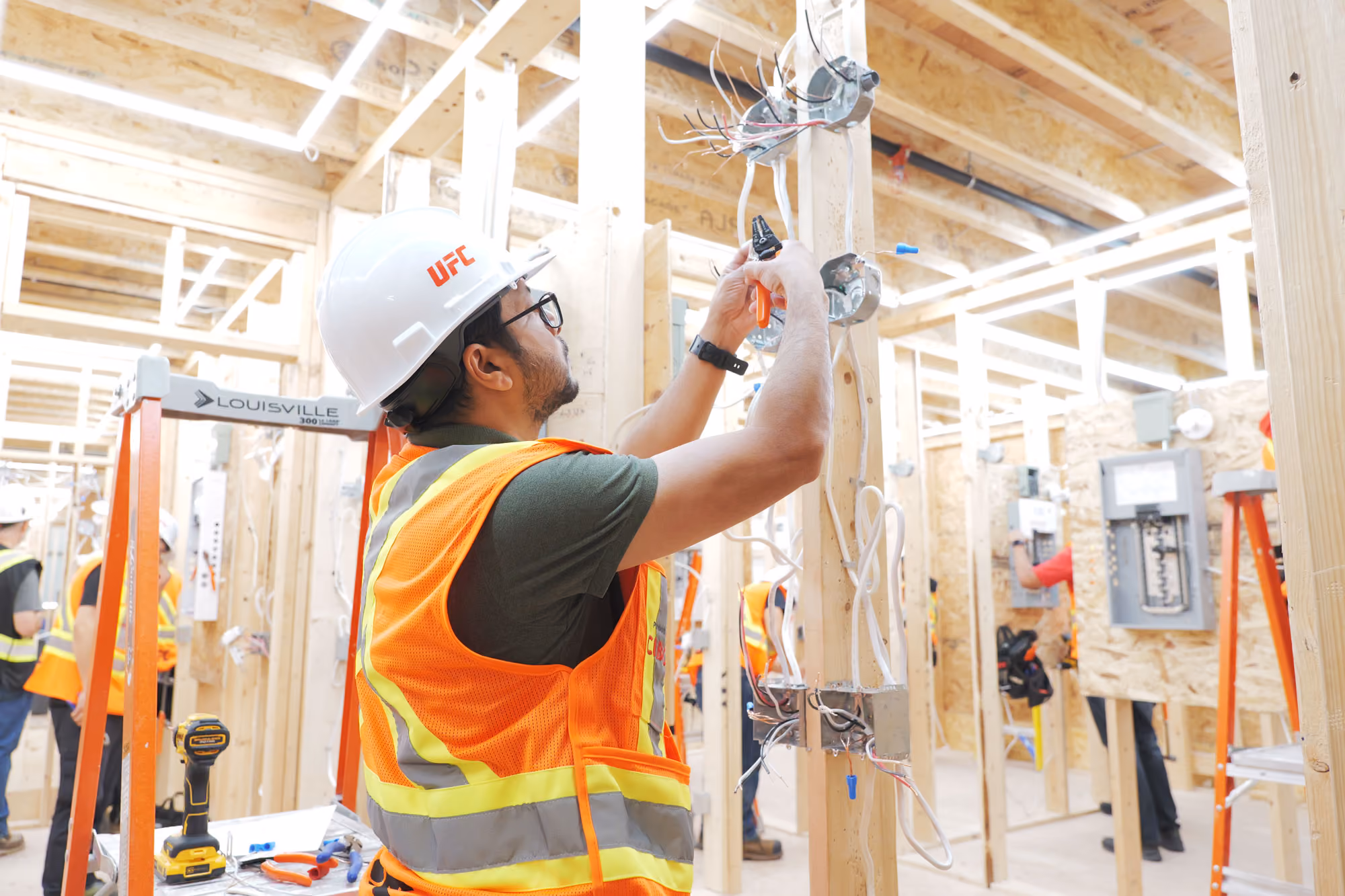 Electrician in a white UFC hard hat and orange safety vest working on wiring attached to a wooden frame inside a construction area with exposed beams and multiple electrical panels.
