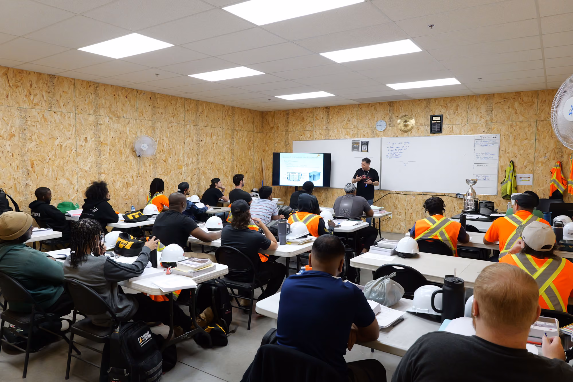 Classroom setting with a diverse group of adult students seated at tables, wearing safety vests and hard hats, facing a male instructor who is presenting technical content displayed on a screen and whiteboard in a wood-paneled room.