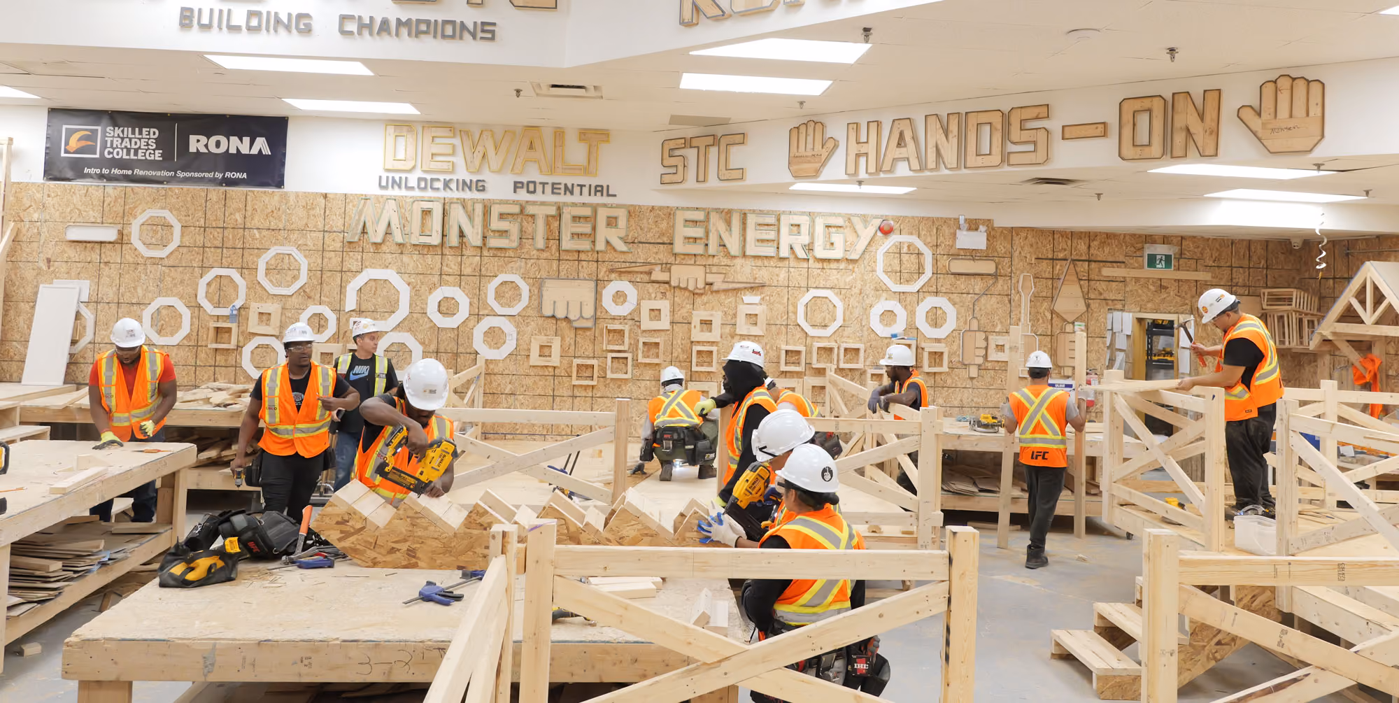 Several people wearing white hard hats and orange safety vests working on wooden construction projects indoors at Skilled Trades College, with wall signs displaying DEWALT, STC, HANDS-ON, MONSTER ENERGY, and building champions.