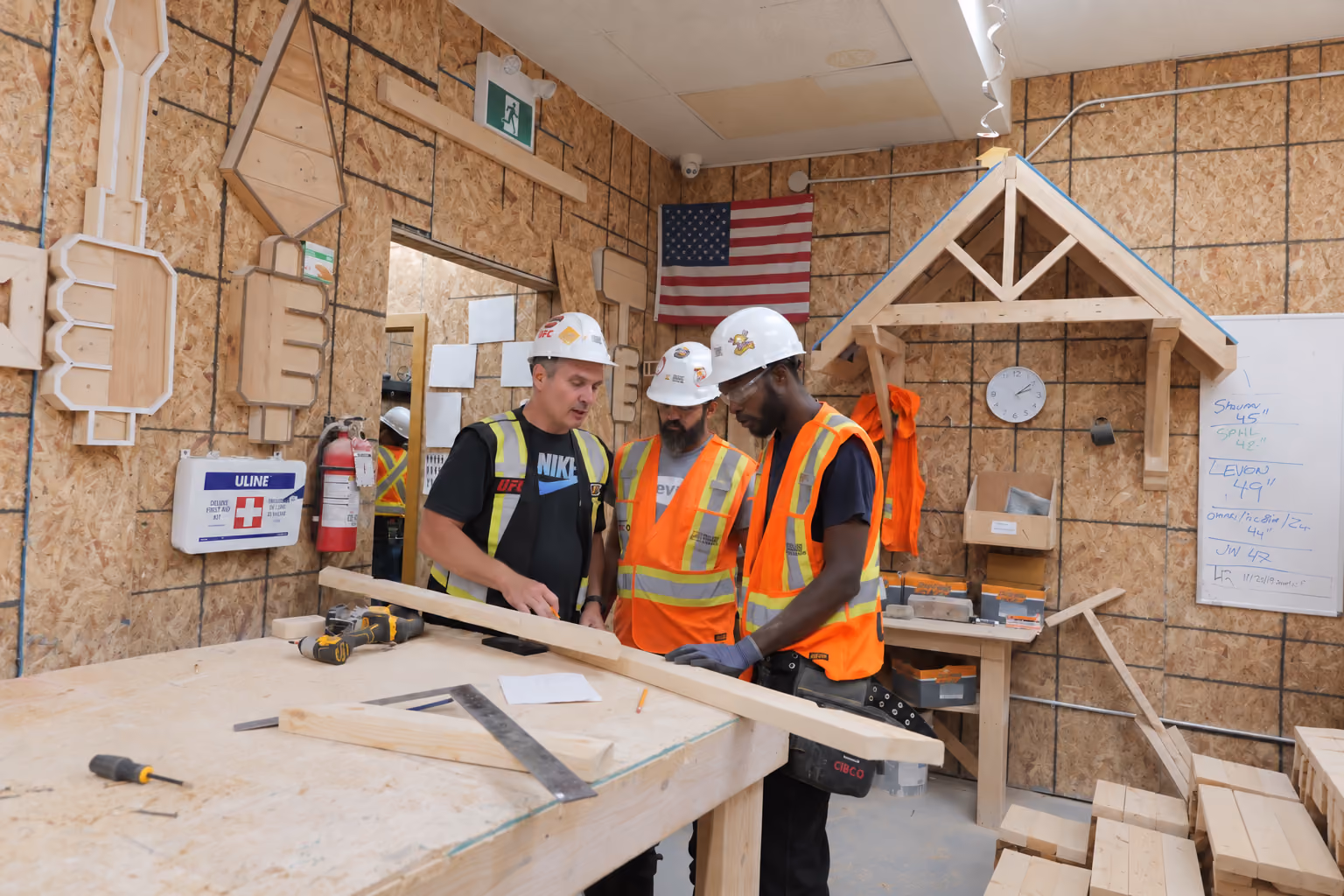Three construction workers wearing hard hats and safety vests examining a wooden beam in a workshop filled with woodworking tools and materials, with an American flag on the wall behind them.