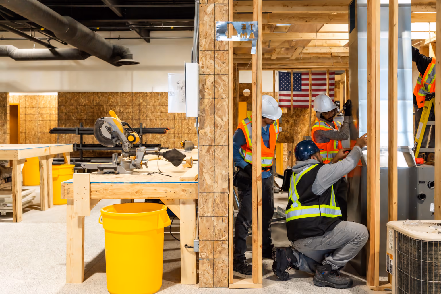 Three construction workers wearing safety vests and helmets installing ductwork inside a wooden frame structure with an American flag visible in the background.