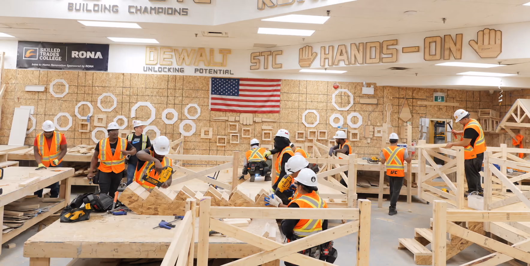 Several people wearing white hard hats and orange safety vests are working on wooden construction projects inside a workshop with signs that read 'DEWALT unlocking potential' and 'STC Hands-On'. An American flag hangs on the back wall decorated with wooden geometric shapes.