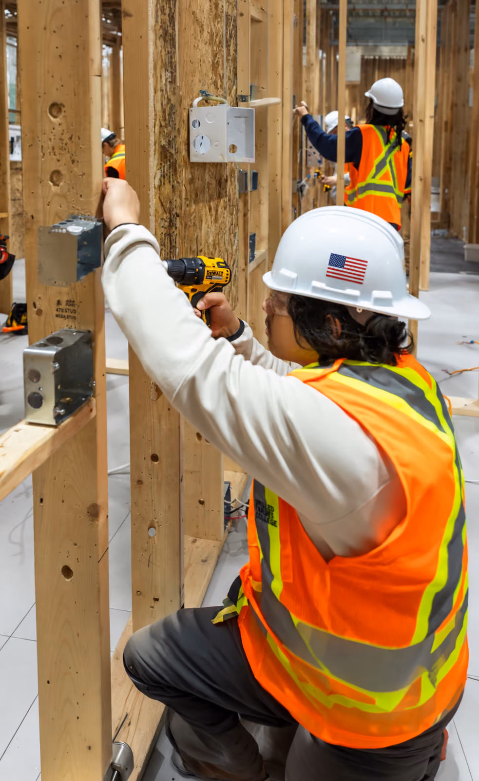 Construction worker in a white hard hat with a U.S. flag sticker and an orange safety vest using a cordless drill on a wooden frame in an indoor construction site, with other workers in safety gear in background.