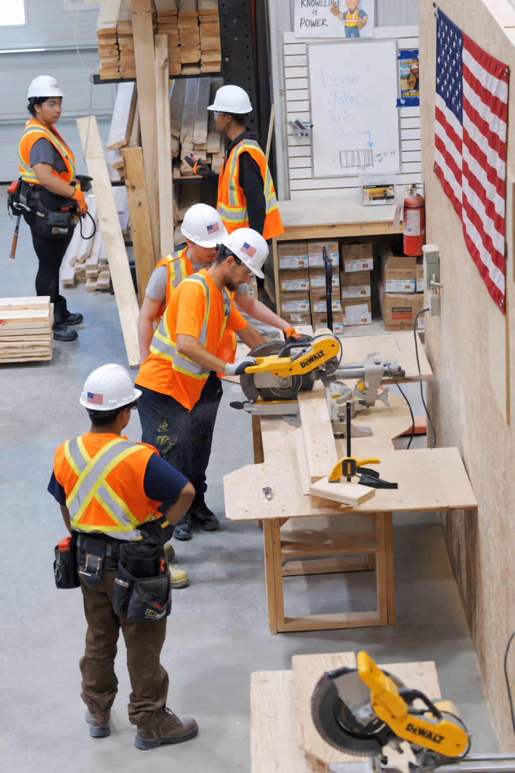 Five construction workers wearing white hard hats and orange safety vests working in a workshop with stacks of wood; one is using a DeWalt miter saw to cut a piece of lumber on a wooden bench, while others watch or prepare materials, with an American flag hanging on the wall and a whiteboard in the background.