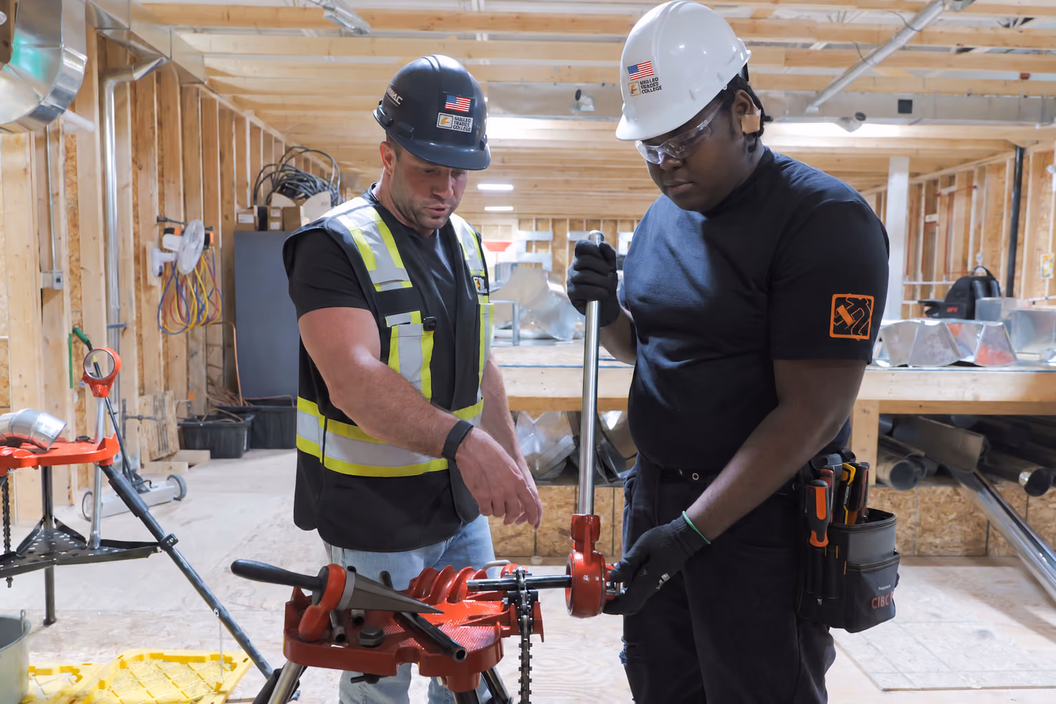 Two construction workers wearing helmets and safety gear are adjusting a large red pipe threading machine inside a building under construction with exposed wooden framing.