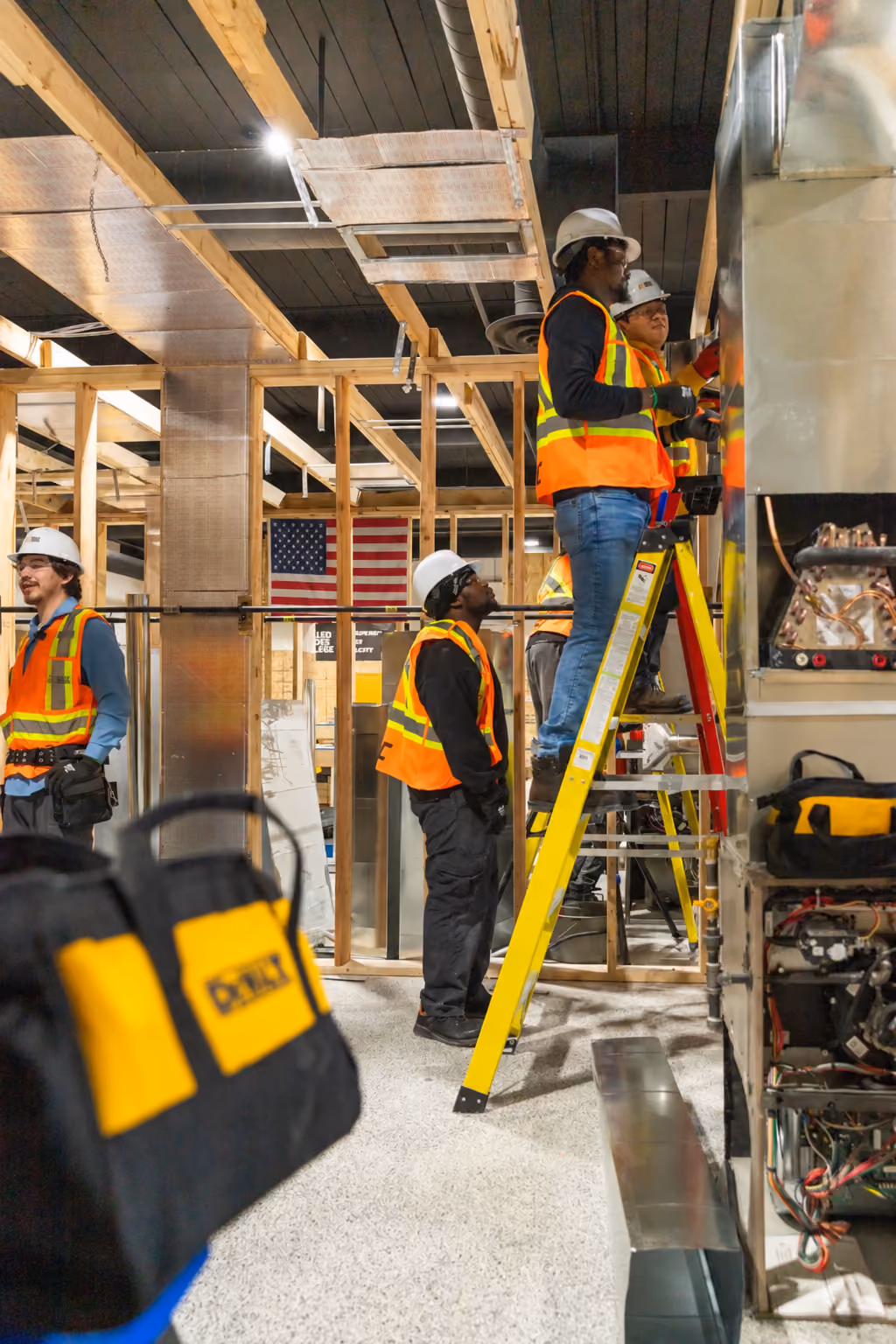 Four construction workers in safety helmets and orange reflective vests working on HVAC ductwork inside a building under construction, one standing on a yellow ladder and adjusting ducts, an American flag hanging in the background.