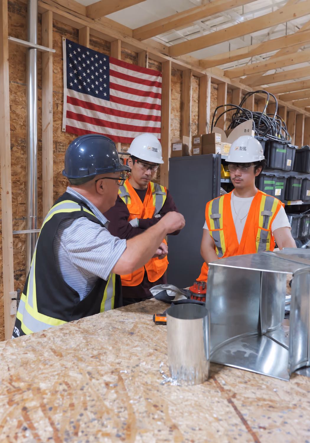 Three construction workers wearing safety helmets and reflective vests inside a wooden framed building under construction, discussing near a table with metal duct components, with an American flag hanging on the wall behind them.