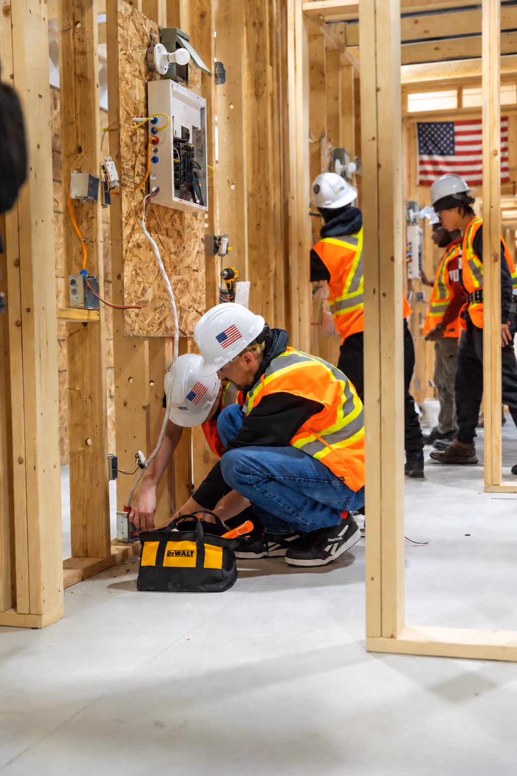 Workers in safety vests and hard hats with American flag stickers install electrical wiring inside a wooden framework in a construction site with a DeWalt tool bag on the floor.