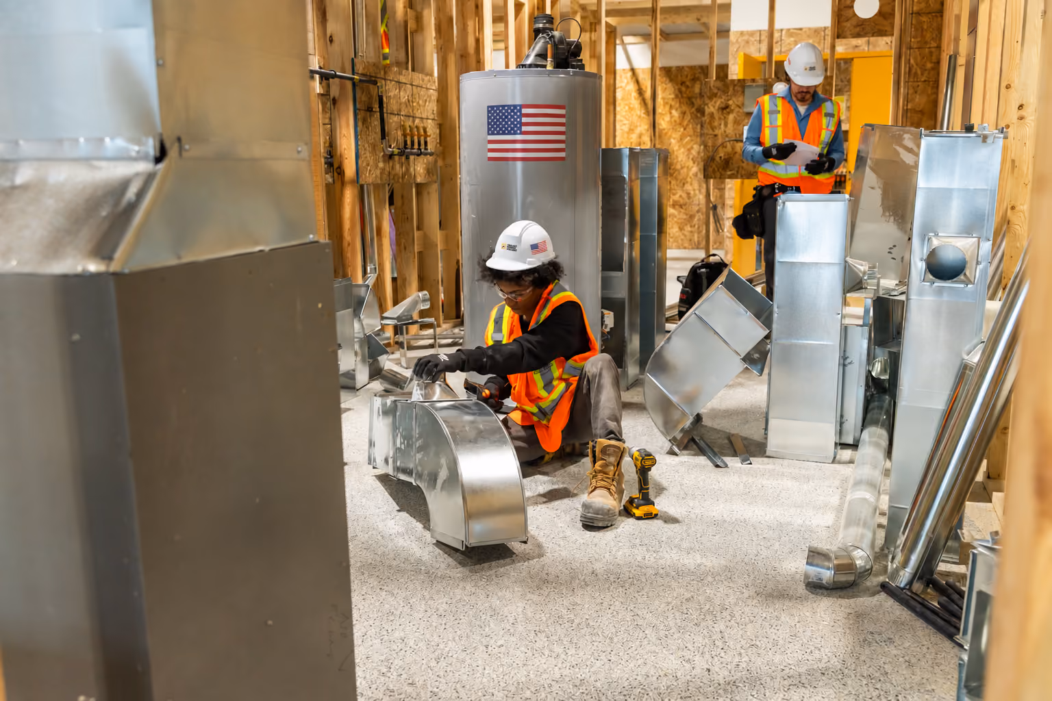 Two construction workers in hard hats and reflective vests working with metal ductwork in an unfinished building room with wooden framing and a large cylindrical water tank with an American flag decal.