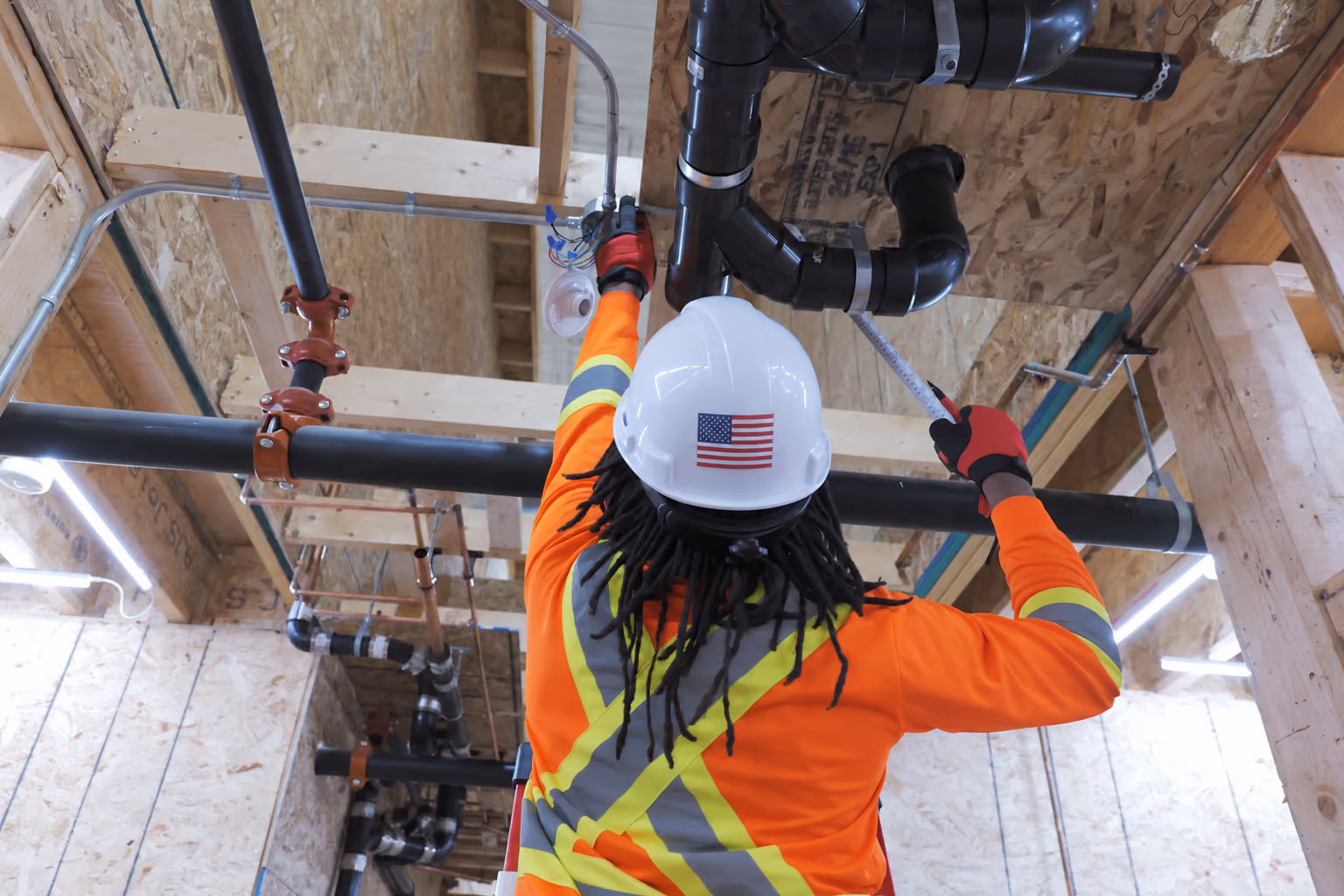 Worker in an orange reflective jacket and white hard hat with an American flag sticker measuring black pipes on a wooden ceiling in a construction area.