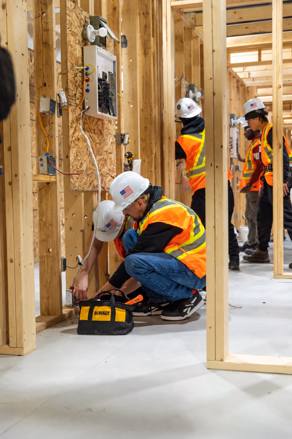 Construction workers wearing hard hats with American flag stickers and reflective safety vests working inside a wooden-framed building under construction, with one worker crouching near electrical wiring and a DeWALT tool bag on the floor.