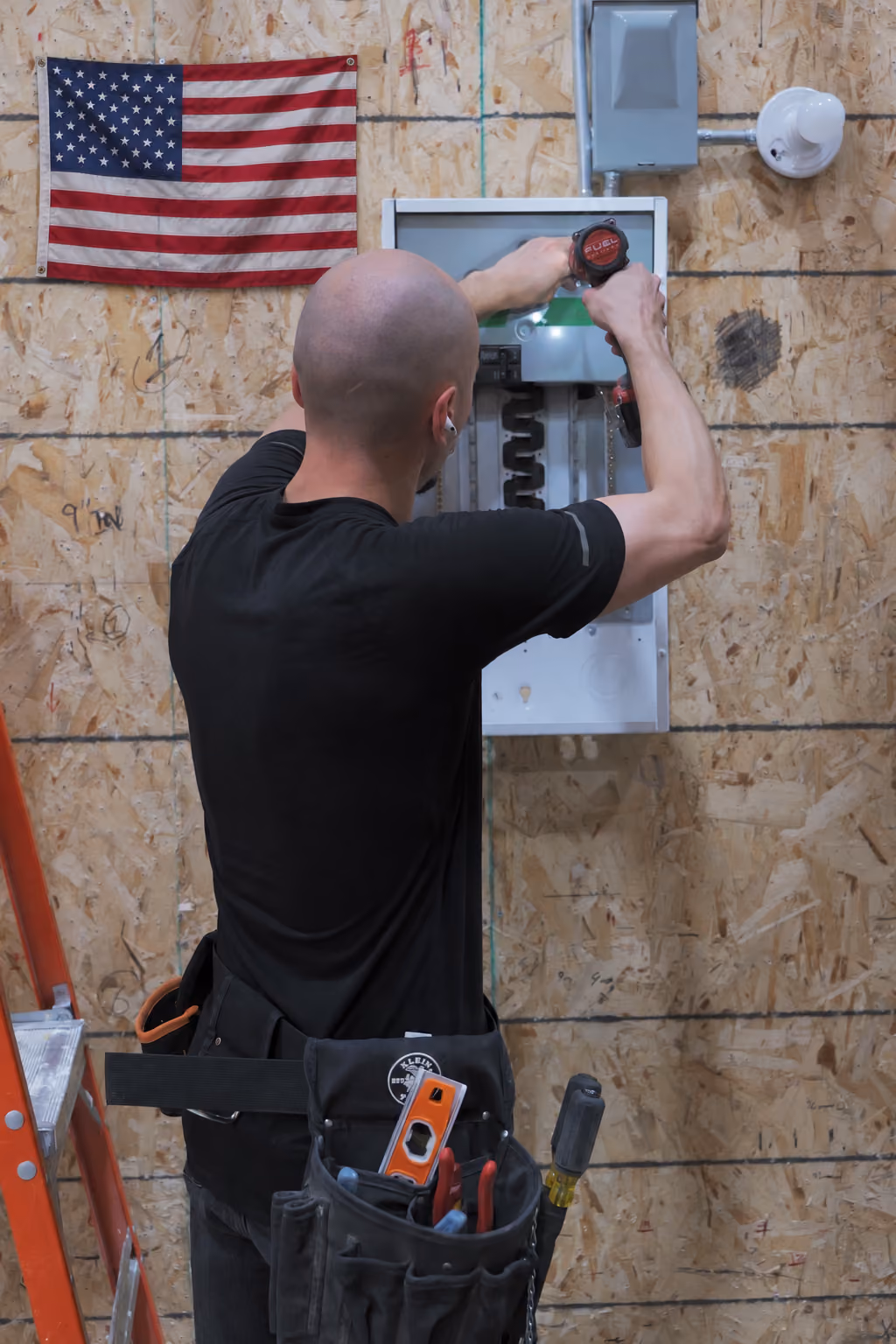 A bald man wearing a black shirt uses a power drill to work on an open electrical panel mounted on a plywood wall with an American flag hanging above. He has a tool belt with various tools and a ladder nearby.