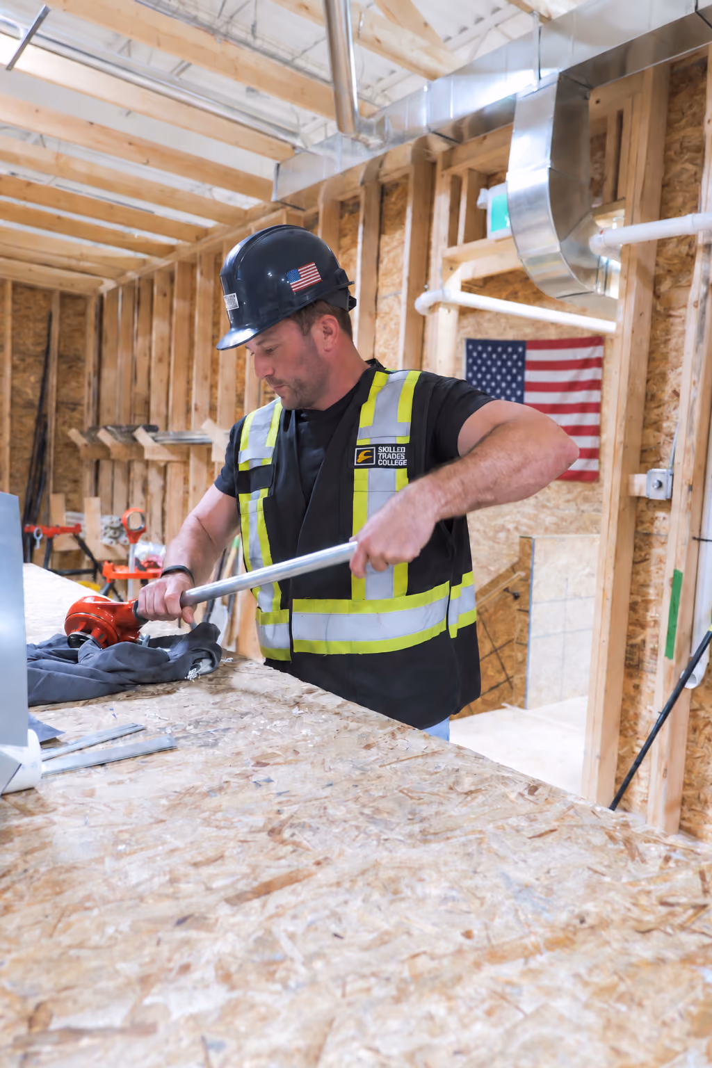 Construction worker wearing a black hard hat with an American flag sticker and a yellow reflective safety vest working with a pipe wrench on a wooden workbench inside a building under construction with exposed wooden beams and an American flag hanging on the wall.
