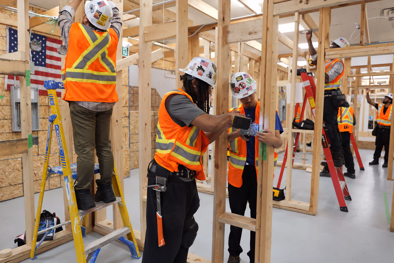 Five construction workers wearing white hard hats and orange safety vests with reflective stripes, working inside a wooden framed building under construction, with two workers on ladders and others securing electrical boxes and wiring to wooden studs.