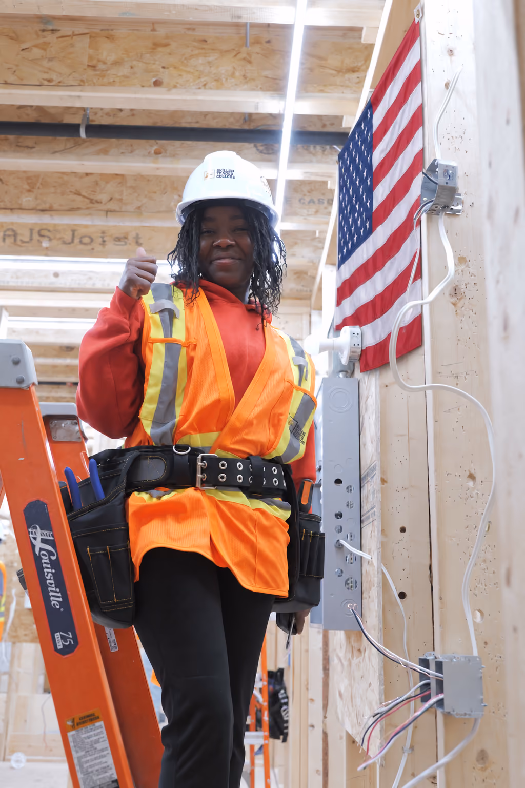 A woman construction worker wearing a white hard hat, an orange safety vest with reflective stripes, and a tool belt stands on an orange ladder inside a wooden frame structure next to an American flag mounted on the wall.